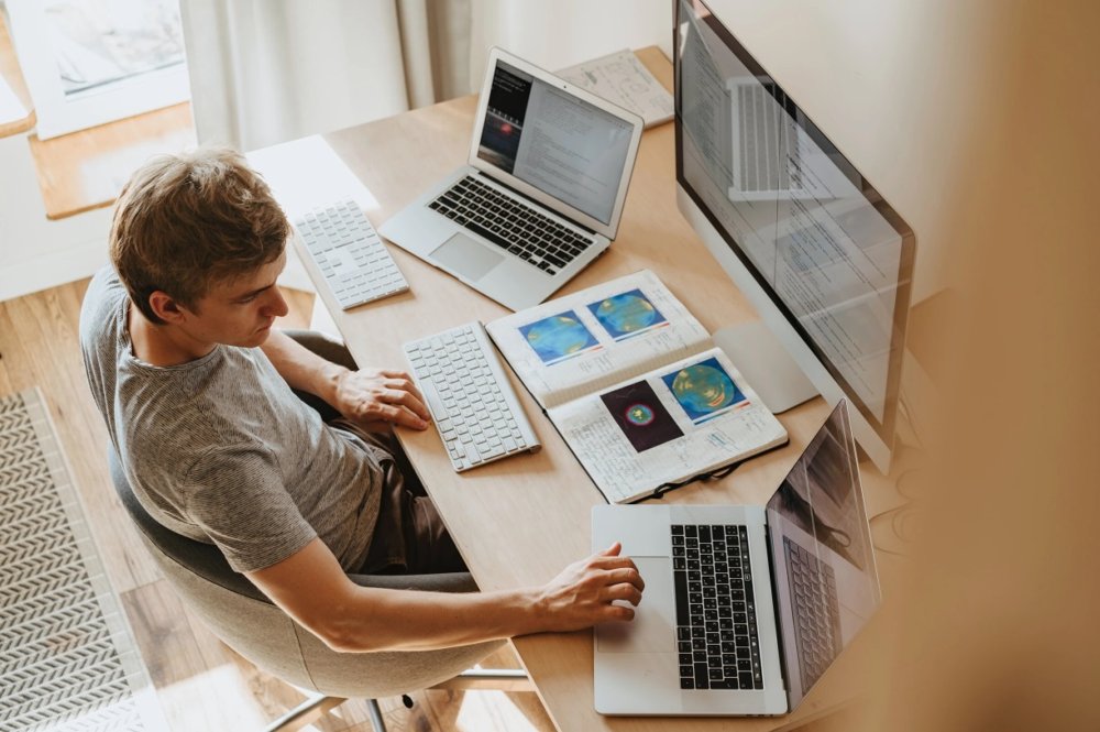 A man sitting at a desk with an open book and two laptops and a large monitor.
