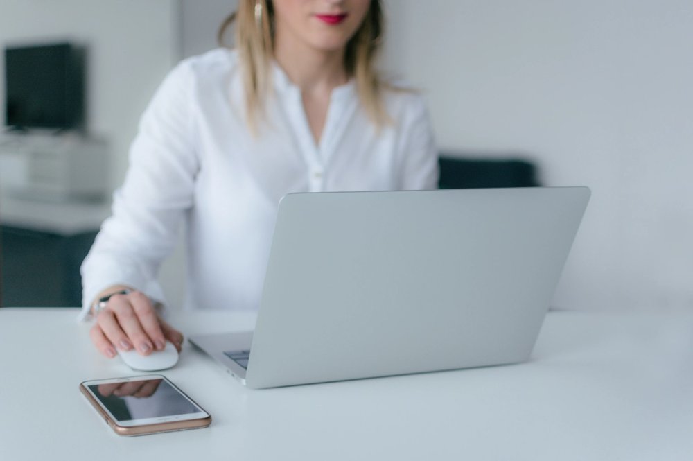A woman in a white blouse sitting sitting at a desk looking at her laptop.